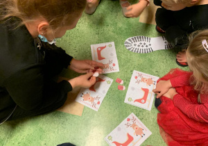 Dzieci siedzą pochylone nad rozłożonymi na podłodze kartkami. Na kartce widać renifery, czerwone sanie Mikołaja, obok widać lad buta.